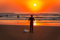 Man in Black Shirt and Shorts Holding White Surfboard Walking on Beach during Sunset