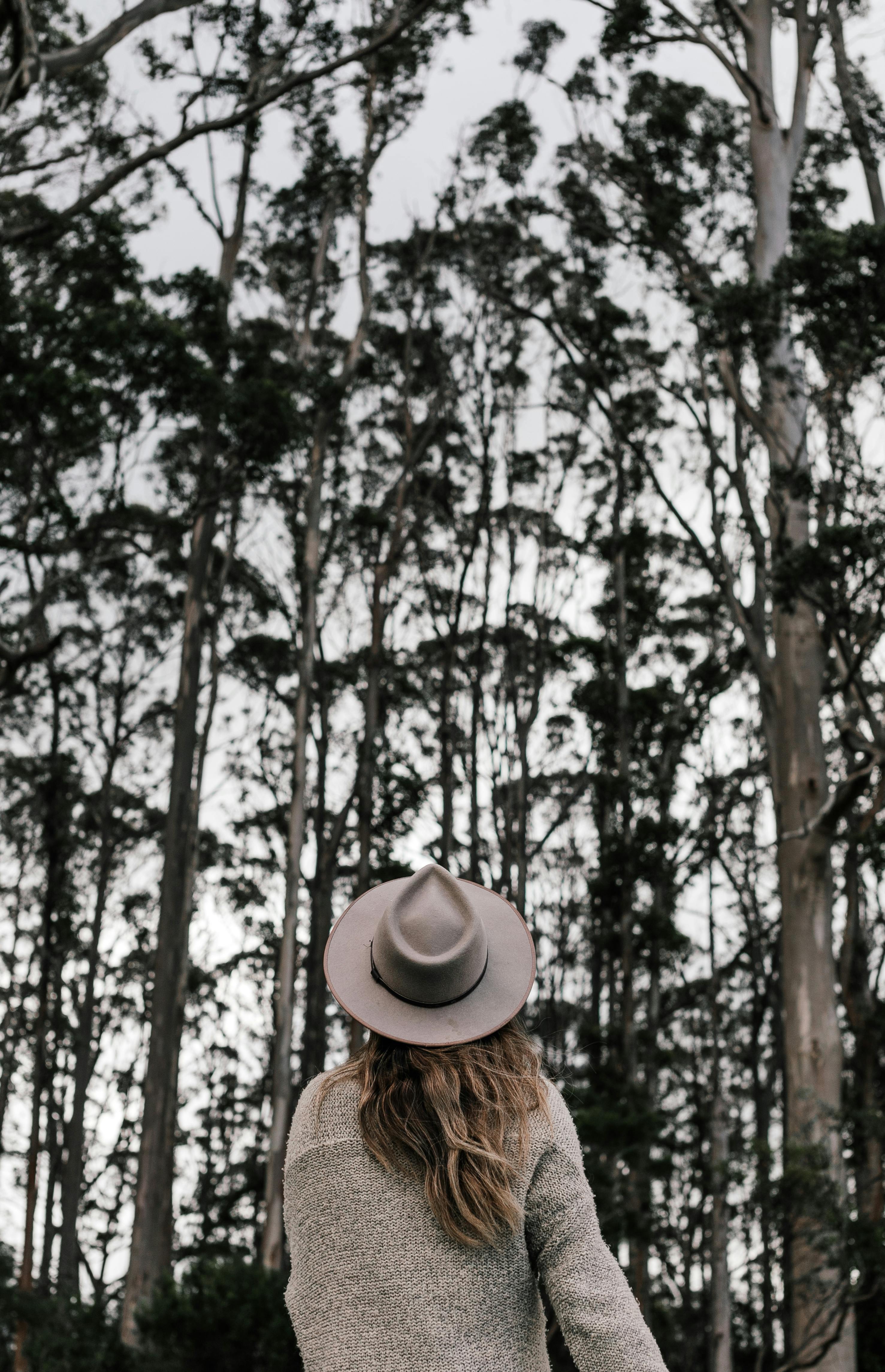 白天，一位身穿时尚服装、头戴帽子的匿名女性背对镜头，站在绿树成荫的森林中