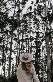 白天，一位身穿时尚服装、头戴帽子的匿名女性背对镜头，站在绿树成荫的森林中