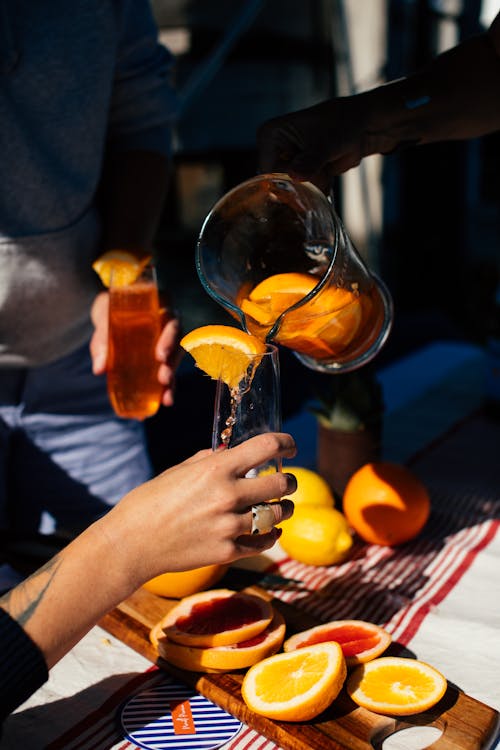 Crop anonymous person pouring fresh cold orange drink from jug into glass during party