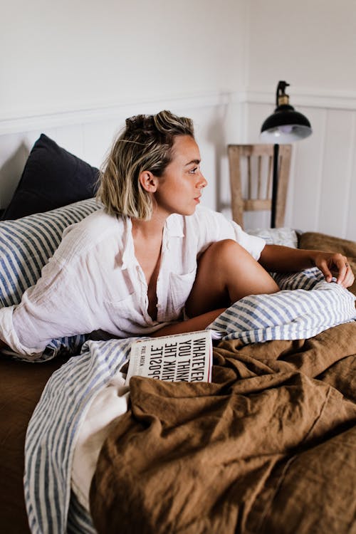 Free Attractive young female in sleepwear sitting on comfortable unmade bed with book and looking away Stock Photo