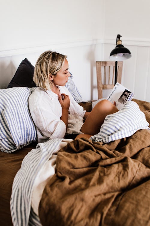 Free Side view of young focused female in sleepwear sitting on comfortable unmade bed and reading interesting book Stock Photo