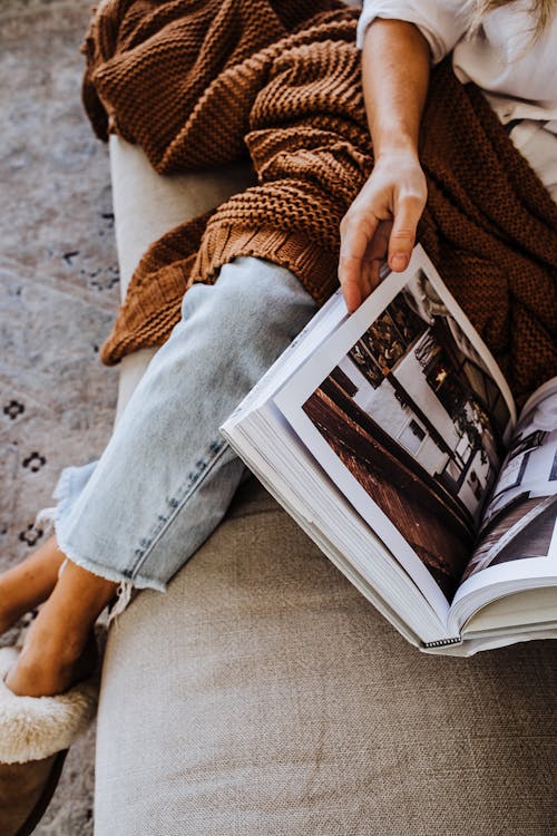 From above of crop anonymous female in jeans sitting on couch and turning pages of book with pictures of interior designs