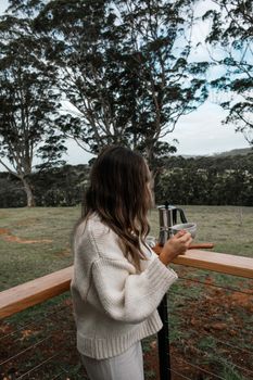 一名穿着休闲装的女子在户外甲板上，一边喝咖啡一边欣赏自然森林景观
