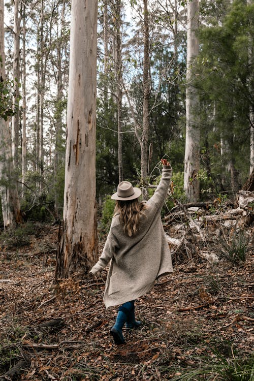 Back view of full body anonymous female in hat and coat walking in woods with raised hand