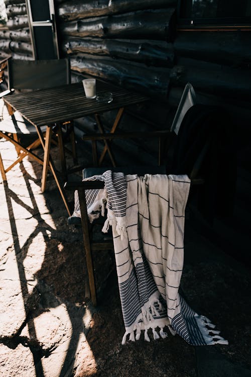 Light outdoor table with chairs and blanket beside wooden house in sunny summer day