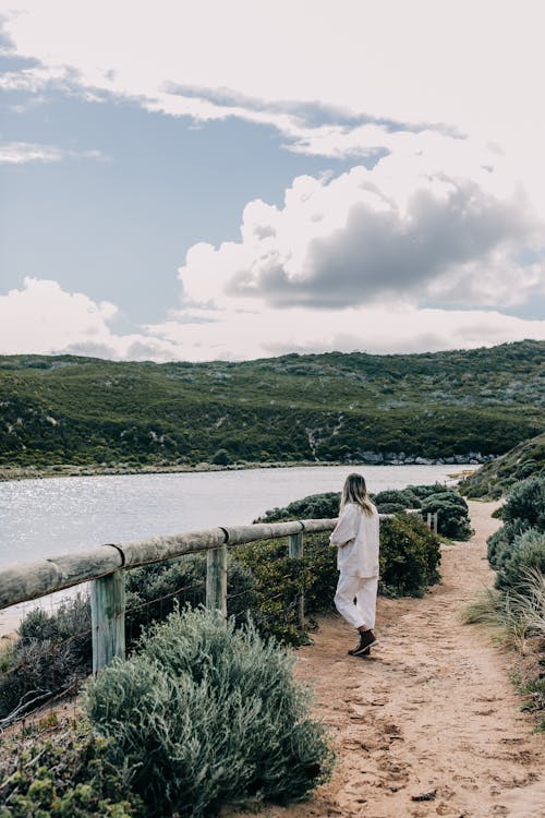 Back view of young female in white casual clothes standing on footpath along river in rural place