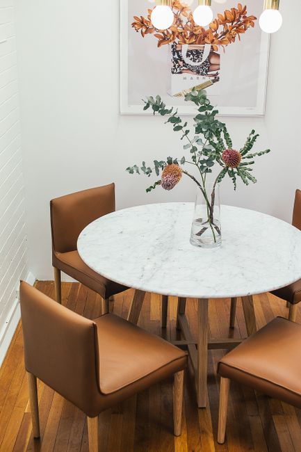 Round table with vase of plants near chairs and white wall decorated with painting