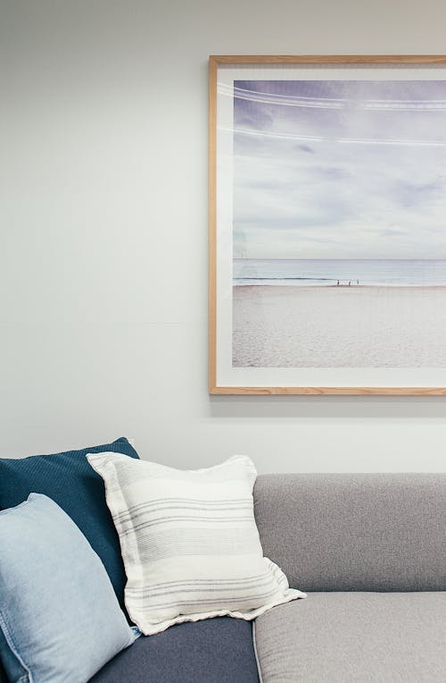 Soft couch with pillows in living room decorated with framed picture of beach and sea hanging on white wall