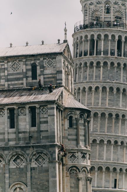Exterior of aged stone church decorated with ornate elements and Leaning Tower of Pisa with tourists on top