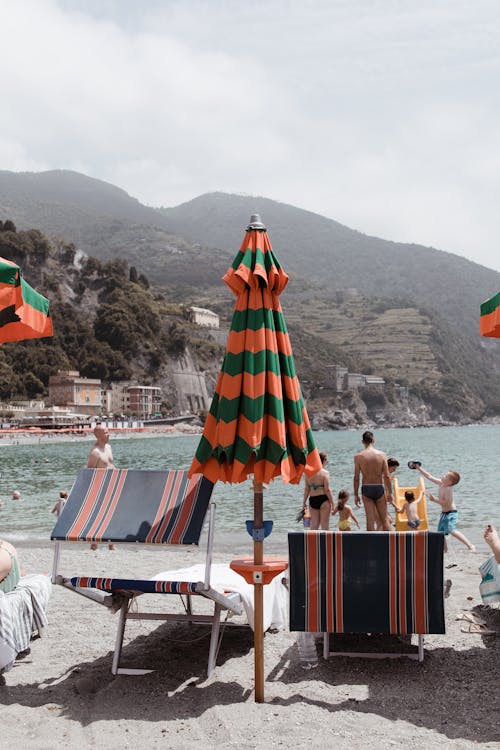 Closed colorful parasol with stripes with sunbeds on sandy beach with people sunbathing and swimming in sea water