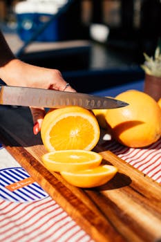 A hand cuts fresh oranges on a wooden board outdoors, capturing a vibrant summer vibe.