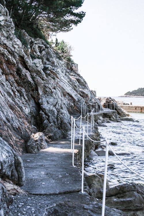 Scenic view of empty fenced footpath between rough mountain and rippled ocean under white sky