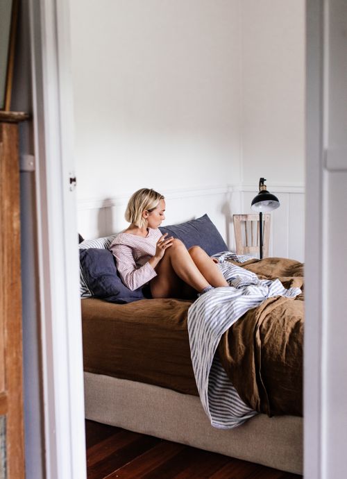 Free Side view of young female sitting on bed with crumpled cover and pillows while looking away in house Stock Photo