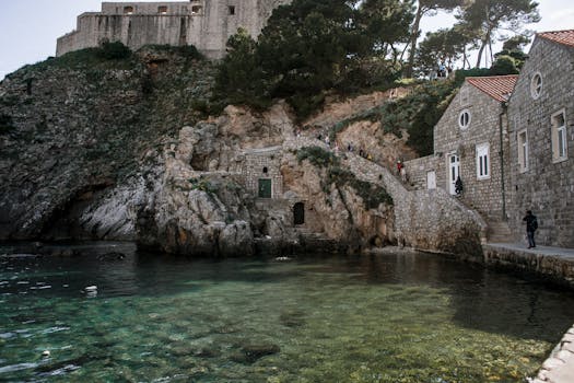 Back view of anonymous travelers on pathway between ancient construction on mount and Adriatic Sea with transparent water in Croatia