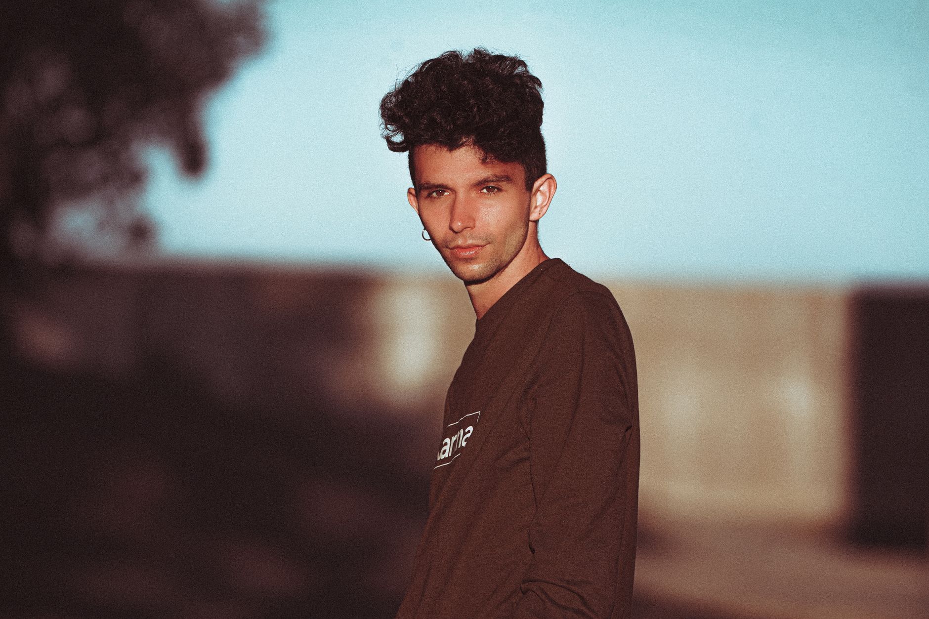 A striking portrait of a man with curly hair and earrings, set outdoors with a vivid background.