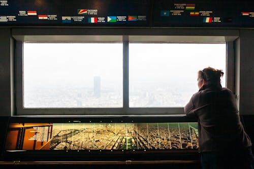 Back view of unrecognizable female traveler standing near window in museum and admiring city on cloudy day