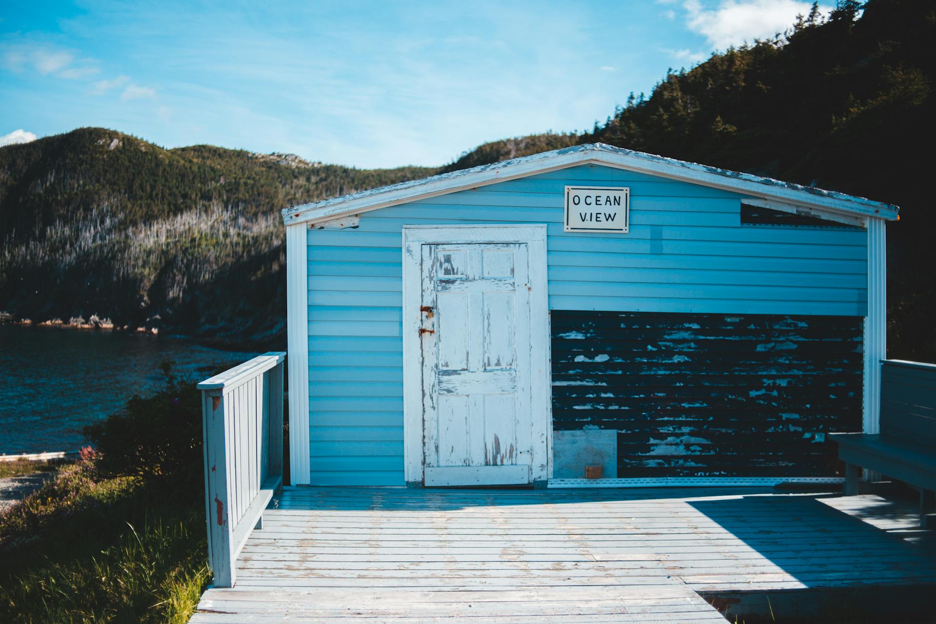 Small old house with shabby door and wall sign with bench placed on terrace on shore of lake in wooded mountains on blue sky