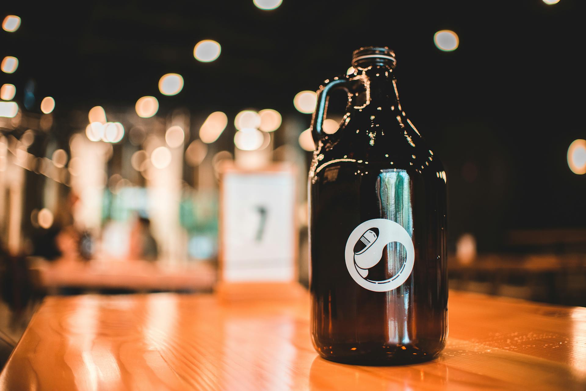 Fancy beer bottle with handle and label with glare on side placed on wooden table in illuminated restaurant