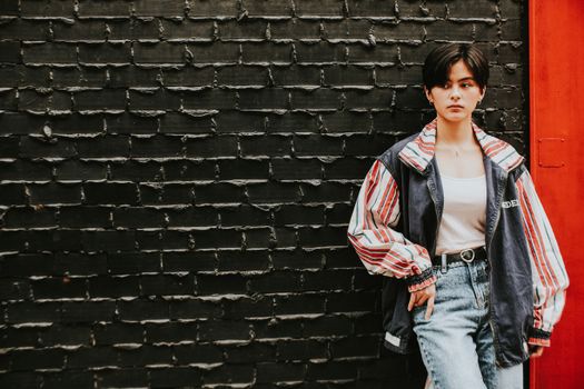一位穿着时尚牛仔布夹克的自信年轻女子在户外黑色砖墙前摆姿势