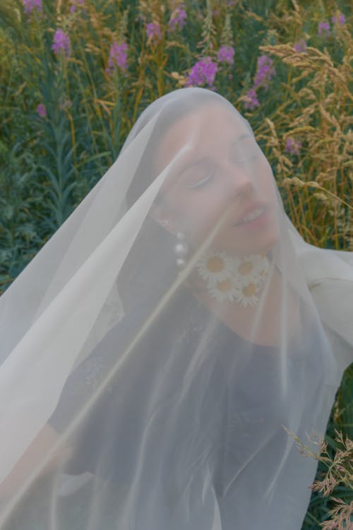 Free Female in earrings with chamomile on neck covered with transparent veil in herbal meadow in sunny afternoon Stock Photo