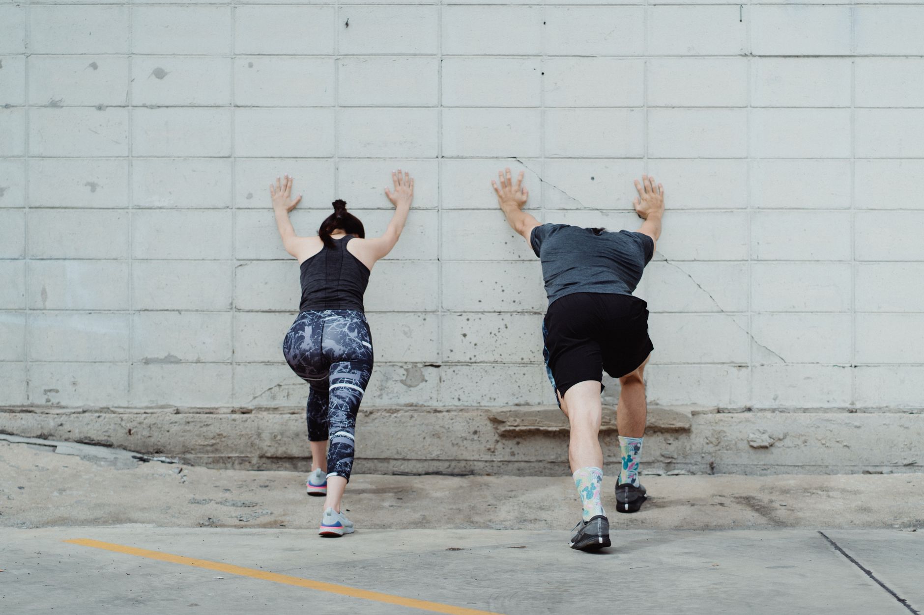 A man and woman in sportswear stretching against a wall for exercise outdoors.
