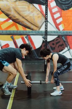 A man and woman working together to lift a barbell in an indoor gym with colorful mural artwork.