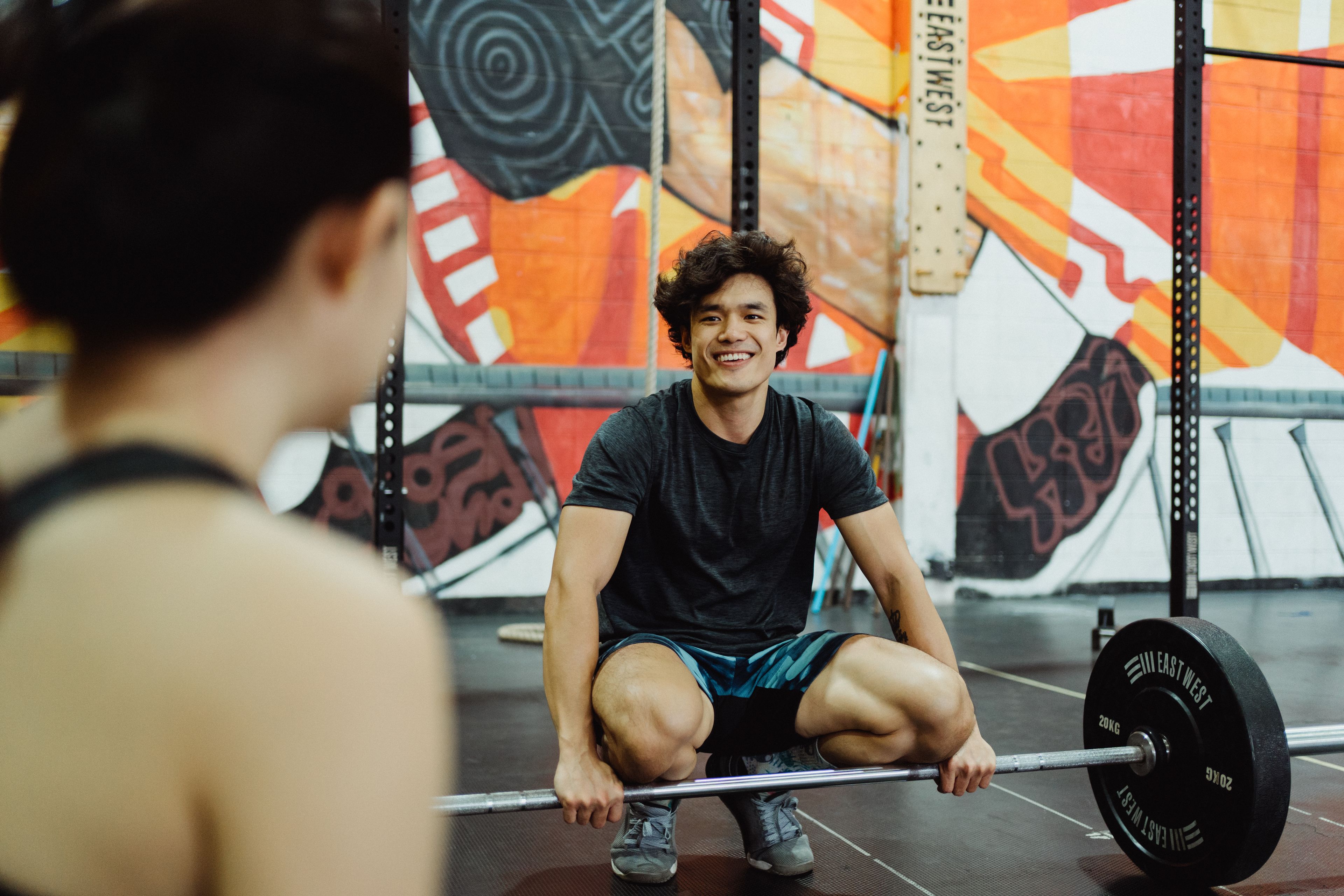 Asian man smiling while squatting with a barbell in a colorful indoor gym setting.