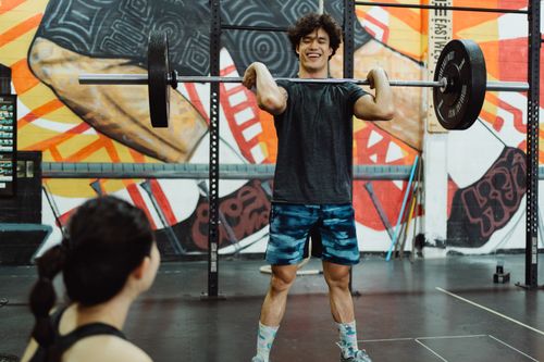Free Asian man performing weightlifting with barbells in front of a colorful gym mural. Stock Photo
