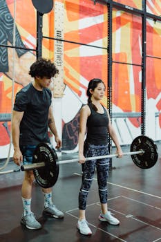 A young man and woman engage in weightlifting exercises at a colorful indoor gym.