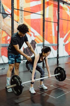 A personal trainer helps a woman with her deadlift form in a colorful gym.