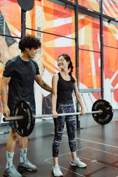 A man and woman smiling while lifting weights in a colorful gym. Fitness and camaraderie.