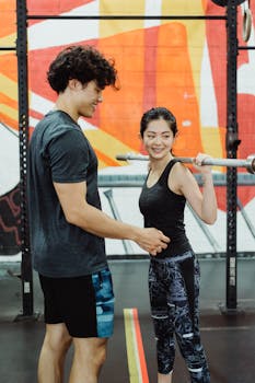A man and woman performing barbell exercises together in a colorful gym.
