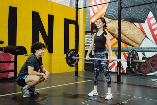 A man assists a woman lifting weights in a vibrant gym environment.