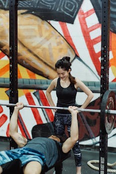 Man performing bench press exercise with woman spotter in a colorful gym setting.
