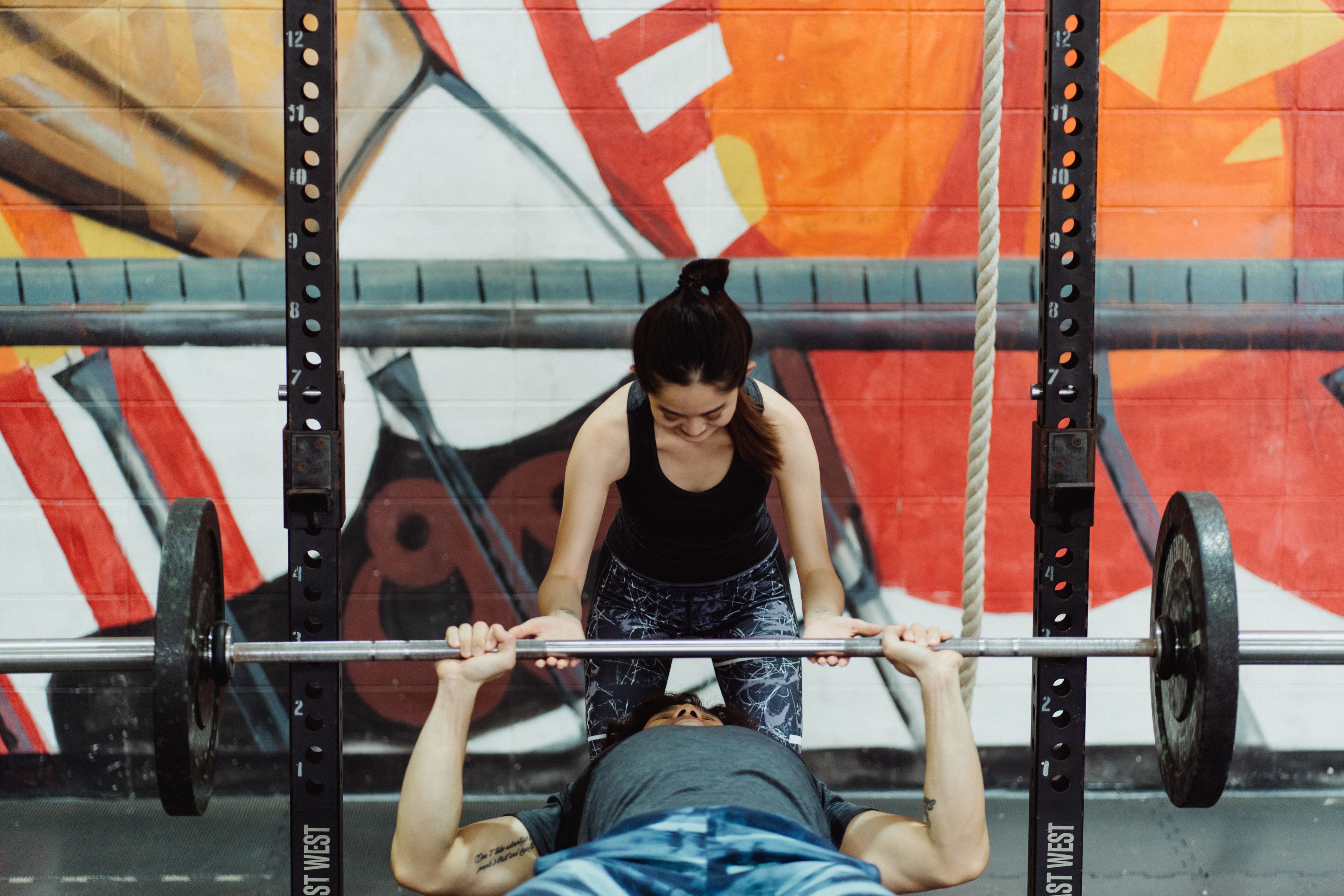 A woman assists a man in bench pressing at a gym with vibrant graffiti walls.
