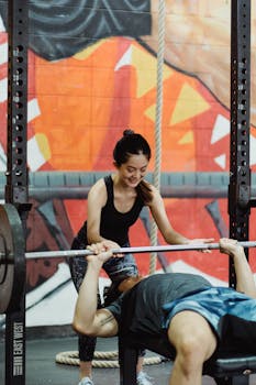 Asian woman assists man during a bench press workout session in a colorful gym.