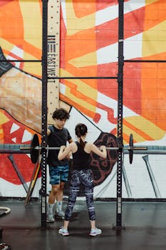 Two adults lifting weights in a vibrant gym with a mural background.