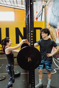 A man and a woman share a fist bump in an energetic gym setting, surrounded by fitness equipment.