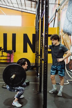 Woman squatting with barbell under supervision in an indoor gym setting.