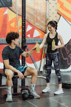 An Asian couple in activewear exchanging drinks in a gym setting, smiling and happy.