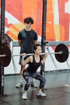 Asian woman squatting with a barbell under trainer's guidance in a vibrant gym setting.