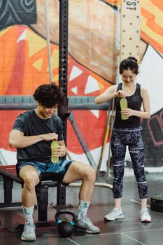 Asian couple in activewear enjoying drinks after a workout in a vibrant gym setting.