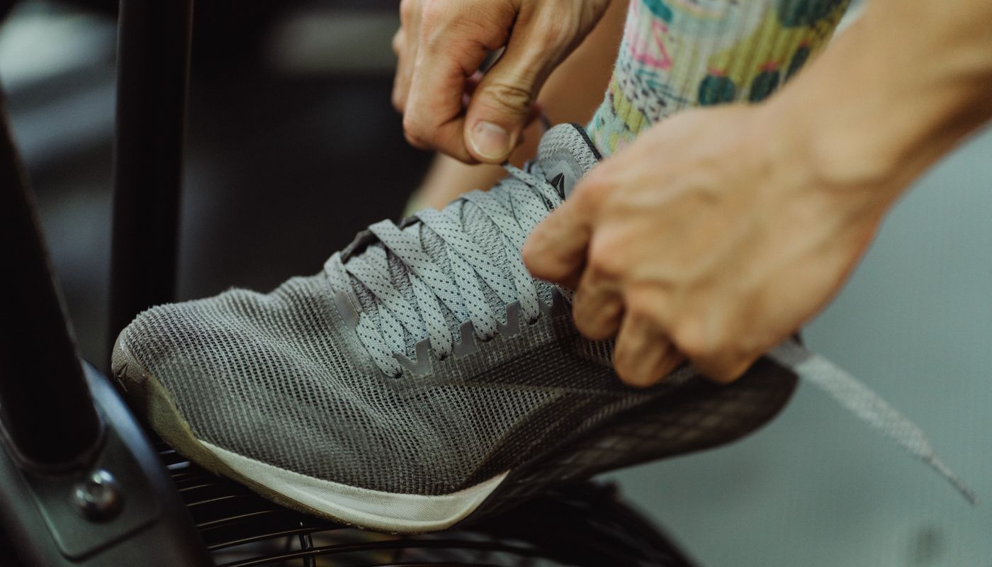 Close-up of two hands pulling tight the laces of a worn grey training shoe resting on a gym machine footplate