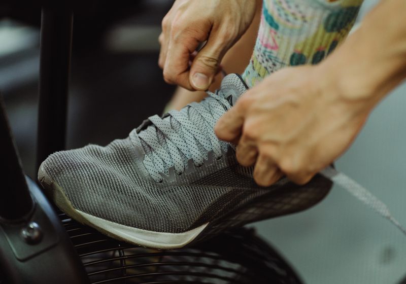 Close-up of two hands pulling tight the laces of a worn grey training shoe resting on a gym machine footplate