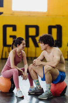 Asian couple in activewear resting on medicine balls in a gym setting, enjoying a moment of relaxation.
