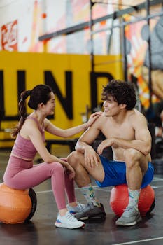 A happy couple in activewear rests on medicine balls in a vibrant gym environment.