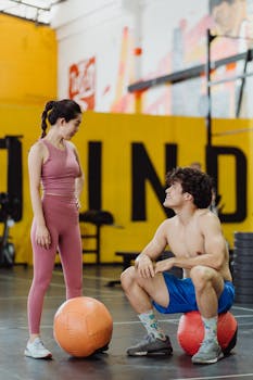 A shirtless man and fit woman interacting with medicine balls in a vibrant gym setting.