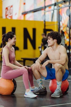 Smiling couple in gym attire taking a break on medicine balls, creating a lively fitness atmosphere.