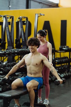 A man performs dumbbell exercises with guidance from a personal trainer in a gym setting.